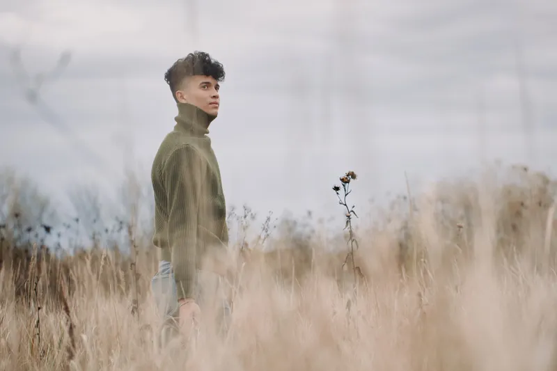 A young man stands in tall dry grass wearing a turtleneck sweater, a contemplative unposed portrait conveying authentic presence and credible emotion in soft natural light and muted earthy tones.