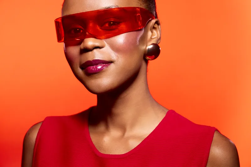Confident black woman with dark skin wears red sleeveless top and futuristic red glasses. She poses against bright orange background, showcasing modern fashion and bold style concept.