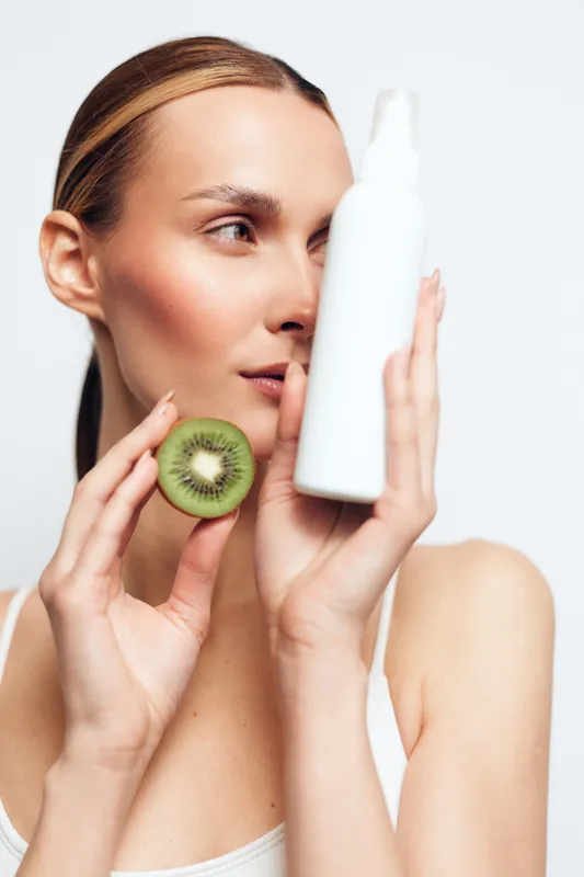 A young woman applies skincare while holding a kiwi slice beside a white cosmetic bottle, conveying a fresh routine and natural beauty in a clean studio setting.