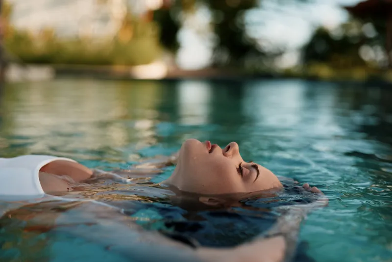 Young woman with brunette hair floats calmly on water surface in a serene outdoor pool during warm daylight, expressing relaxation and peacefulness in a casual white swimsuit.
