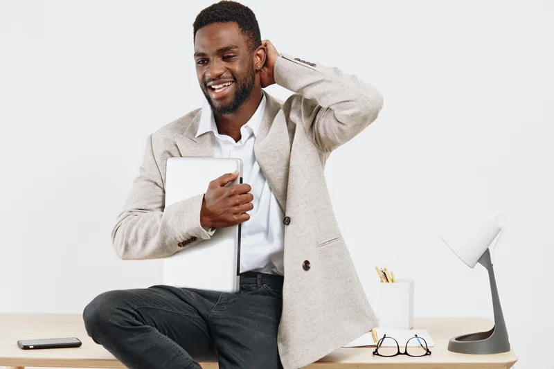Confident smiling man with short curly hair and beard using tablet device while sitting at desk in modern office, casual professional attire, positive mood, light background, workspace environment, bu