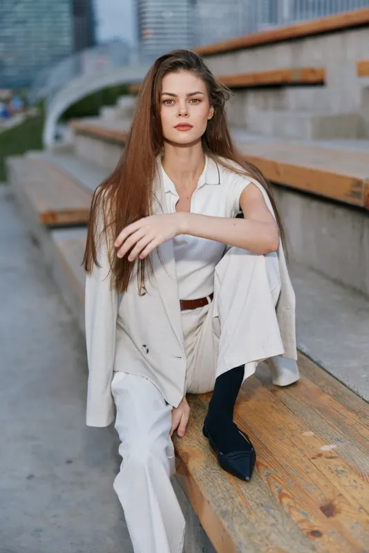 young woman wearing light beige suit and white blouse sitting on wooden urban stairs with confident expression autumn fashion business style