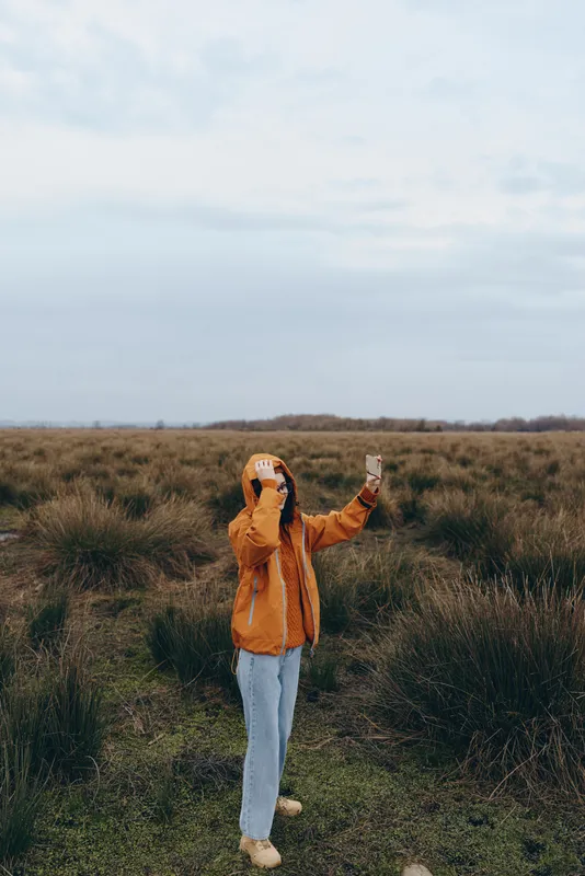 Woman outdoors in a grassy field, orange jacket and white pants, arms raised, looking up at cloudy sky, lifestyle moment, calm stance, soft wind, natural colors