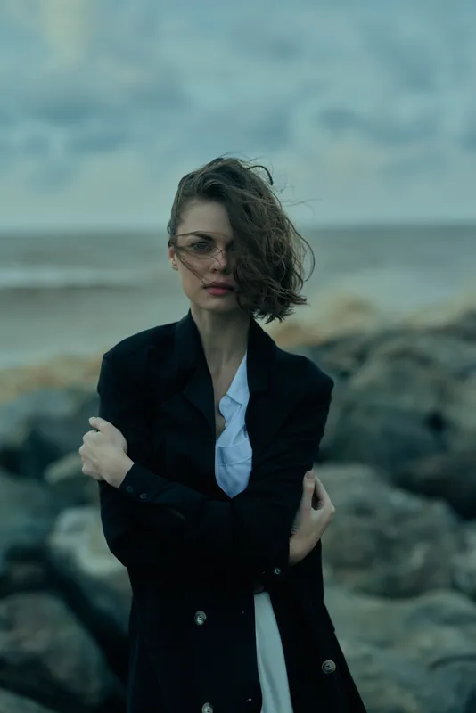 Woman in a dark blazer stands on a rocky coast with wind swept hair, moody ocean backdrop and fashion forward styling for editorial storytelling and dramatic lifestyle imagery