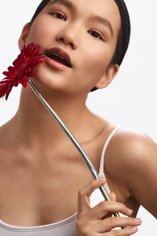 an asian woman holds a red flower close to her lips, posing gracefully in a studio portrait that emphasizes delicate beauty and contemporary elegance.