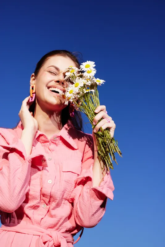 Young woman in a pink dress enjoying a sunny day while holding a bouquet of daisies, smiling against a bright blue sky, conveying happiness and joy outdoors