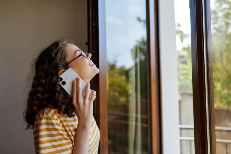 Young woman with curly hair in a yellow striped top speaks on a smartphone by an open window, as bright daylight fills a cozy home and sparks a warm connection.