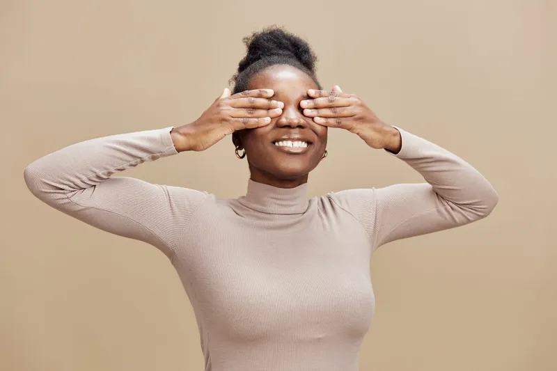 Happy young African woman smiling and playfully covering her eyes with hands against a beige background, showcasing a joyful and carefree emotion