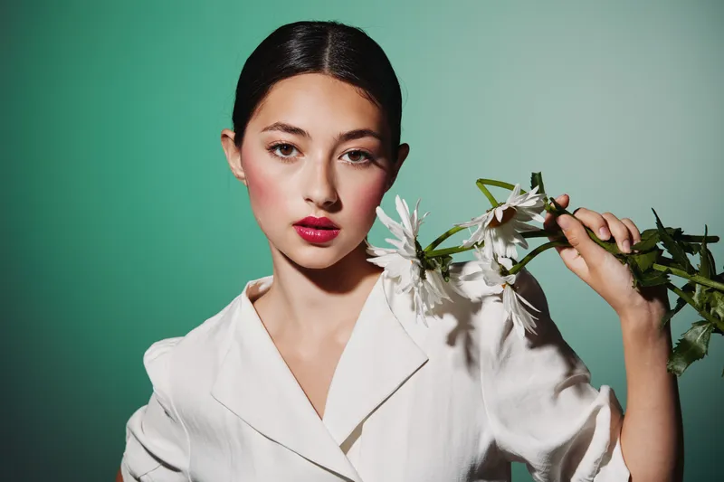 Young model with elegant makeup and slicked hair posing in white outfit holding white daisies. Studio shot on a minimal green background, concept for beauty fashion industry and creative style.
