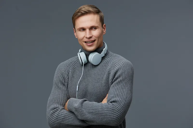 Smiling young man with headphones wearing gray sweater looking away on plain gray background studio portrait. People lifestyle concept, casual, relaxed, male, fashion, technology.