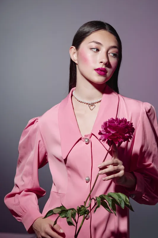 Young woman in a vintage pink jacket holding a dark fuchsia flower close to her body. Soft lighting highlights elegant makeup and sleek straight hair for beauty fashion concept.