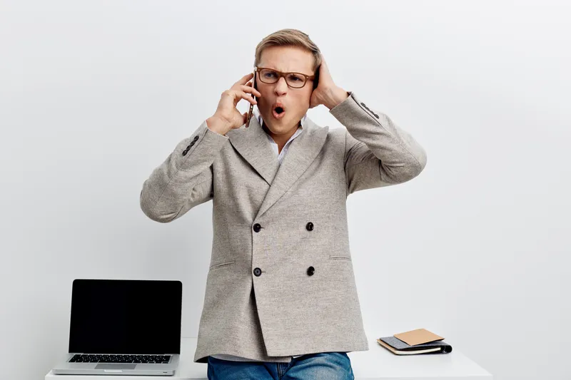 Surprised man wearing glasses and gray blazer standing at desk with open laptop and notebook, reacting to unexpected news or problem, indoor studio shot. Business office concept.
