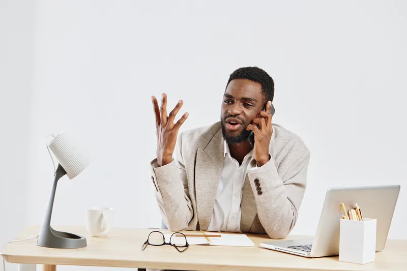 Confident man talking on phone during work at desk with laptop, glasses, pen, and coffee mug in modern office setting. Professional, business, communication, technology, workplace, success.