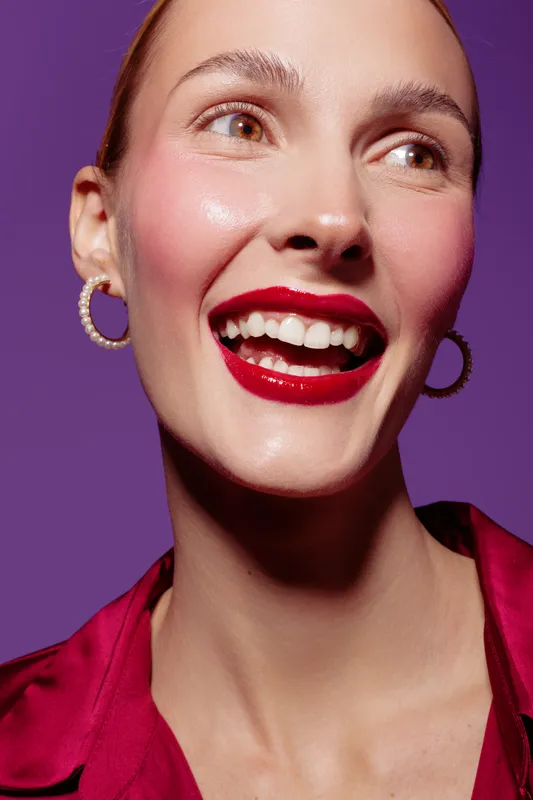 Close-up of a smiling woman with red lipstick and pearl earrings wearing a red blouse. Bright purple background highlights her joyful expression and glowing skin. Beauty and makeup concept.