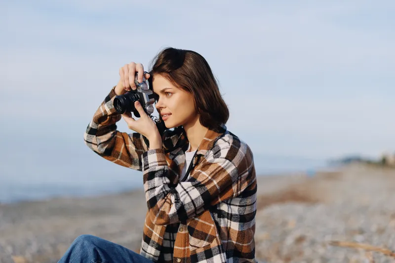 Photographer woman outdoors by the sea using a camera, focused on her shot in a casual plaid jacket, coastal breeze and horizon in view, capturing travel moments with artistic eye