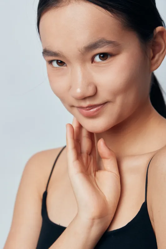 Young woman with flawless skin posing with hand near face in black top on light background. Beauty portrait with natural makeup and confident expression for skincare concept.