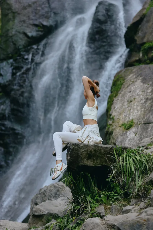 Young woman relaxing near waterfall in white sportswear, sitting on rock surrounded by greenery, enjoying nature and peaceful outdoor scenery.