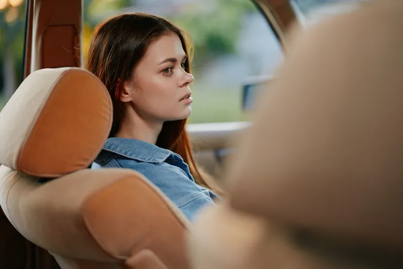 Young woman in a denim jacket looking thoughtfully out of a car window, with an autumn backdrop and soft lighting for a contemplative mood.