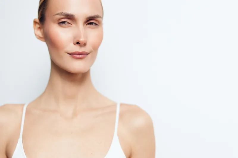 A confident young woman with clear skin and minimal makeup looks directly into the camera against a plain white background. Concept for beauty and skincare.