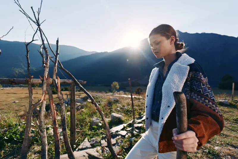 Girl gardening with a spade in a rural farm field at sunrise, wearing a warm jacket and tending soil near wooden stakes with mountains and golden sunlight in the background.