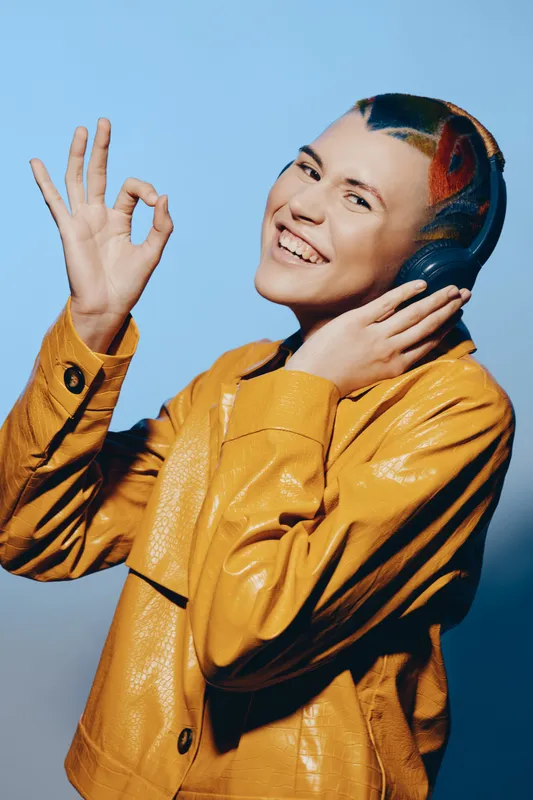 young woman with headphones and colorful hairstyle enjoying music, wearing a stylish yellow jacket and smiling confidently against a blue background