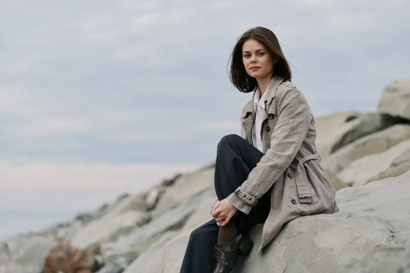 Young woman in a beige coat sitting on rocks by the sea under cloudy sky. Casual outdoor portrait with natural light, relaxed mood, and cool color tones in nature setting.