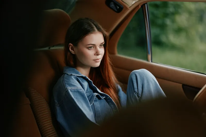 Young woman in casual denim outfit relaxing in a vintage car, showcasing serene emotions amidst a lush green background with natural light.