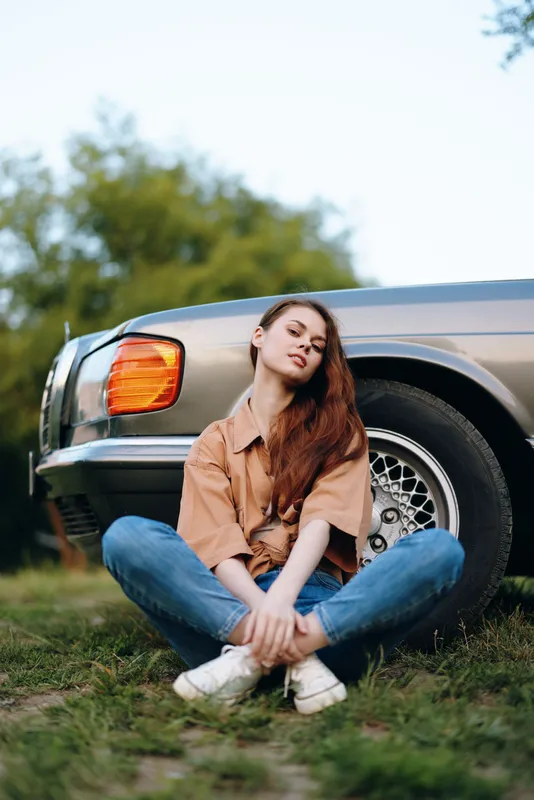 Young woman sitting casually on grass near vintage car, wearing stylish outfit and expressing calm emotions in a natural outdoor setting.