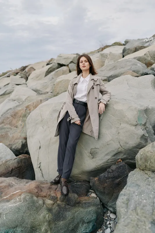 Young woman in beige coat and black pants leans on large rock outdoors with cloudy sky above. Casual fashion outfit in natural rocky landscape. Concept of style and confidence in nature.