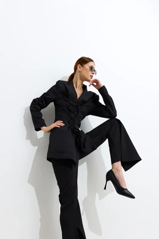 Fashion woman dressed in black suit and heels poses confidently against white wall, wearing sunglasses with relaxed hairstyle, showcasing modern style and attitude.