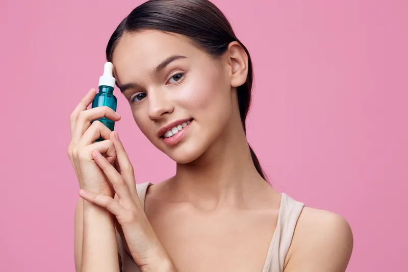 Young woman holding a skincare serum, smiling against a soft pink background, embodying beauty and self care with a fresh, vibrant look