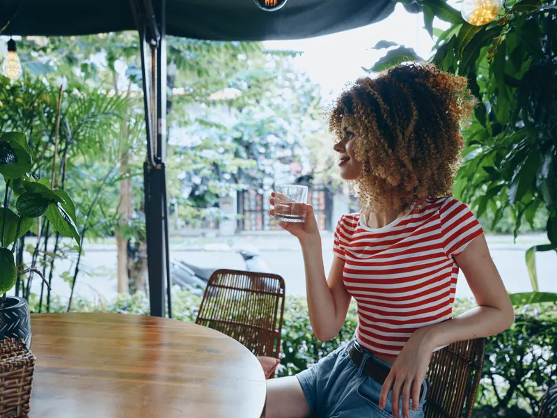 A young woman with curly hair sits at an outdoor cafe table, sipping a drink in a red and white striped tee, framed by lush greenery and warm natural light.