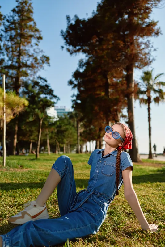relaxed girl wearing blue denim overalls and sunglasses sitting on green grass in sunny park with tall trees and clear sky background