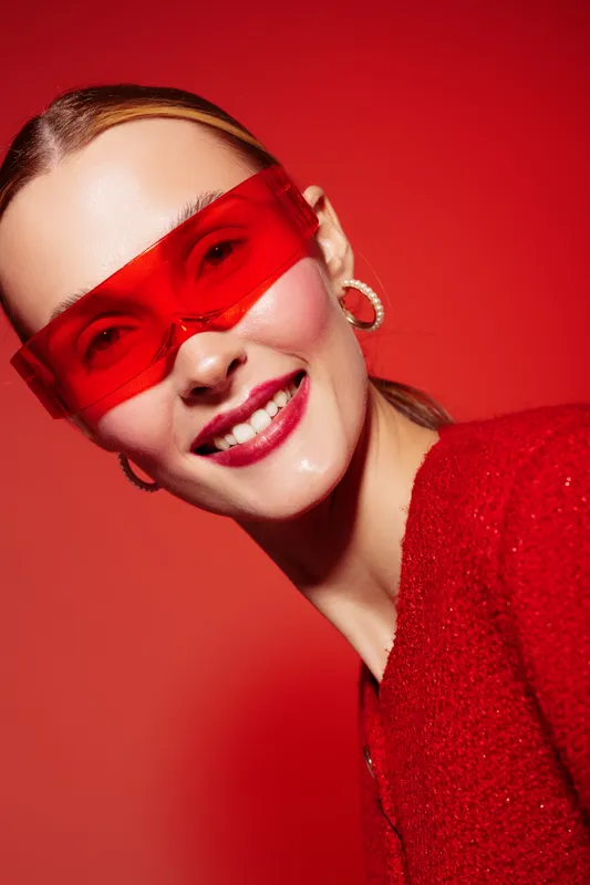 Smiling woman with red sunglasses and red sweater poses against red background. Fashion portrait with bright colors and modern style. Concept of confidence, beauty, and vibrant mood.