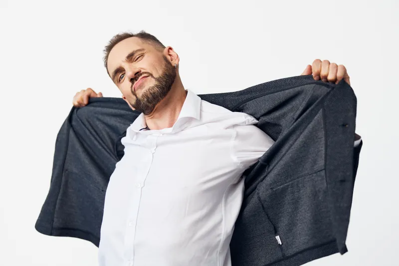A business professional stands relaxed in a white studio background, pulling open a suit jacket to reveal confidence, readiness, and approachable energy.