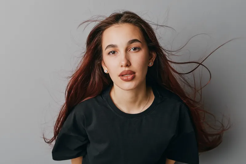 Young woman with long hair wearing black t shirt looking at camera with serious expression on gray background. Portrait of female model with natural makeup and flowing hair indoors.