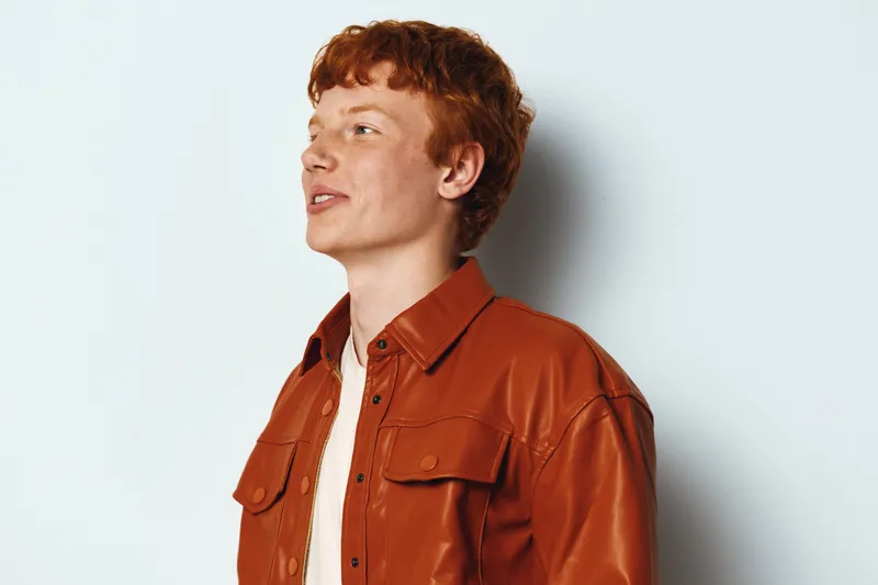 Young man with red hair wearing orange jacket standing against white background looking away and smiling. Casual style, portrait, side view with natural expression and trendy clothing.
