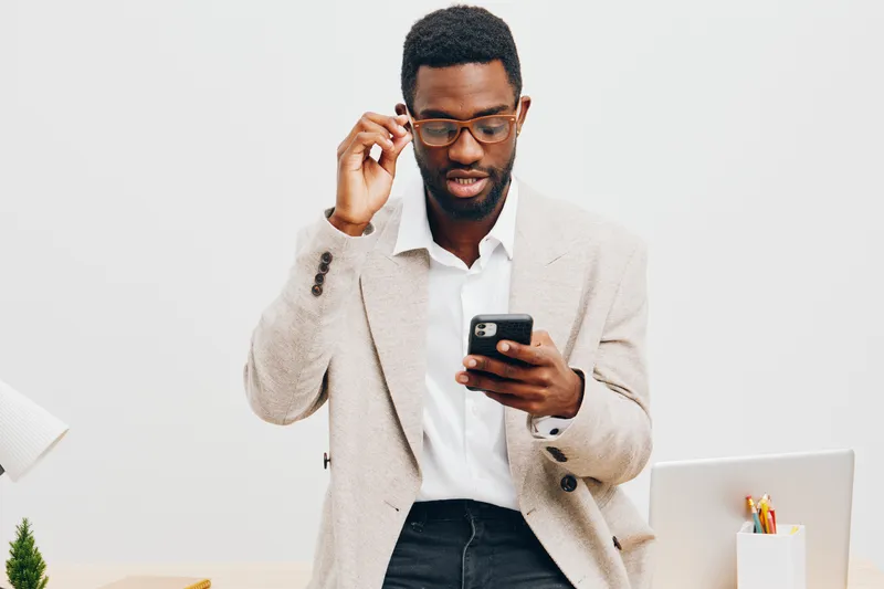Professional young African-American man wearing glasses, white shirt and beige blazer looks surprised while reading message on smartphone, working in modern office with minimalistic design and white b