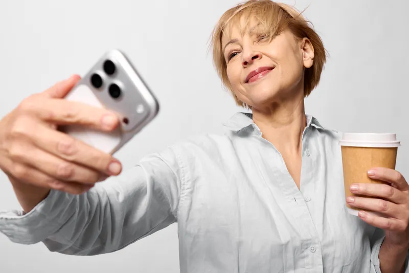 Senior woman holds a smartphone and a coffee mug, smiling toward the screen in a bright white home. Relaxation, communication, online video and calm wellbeing.