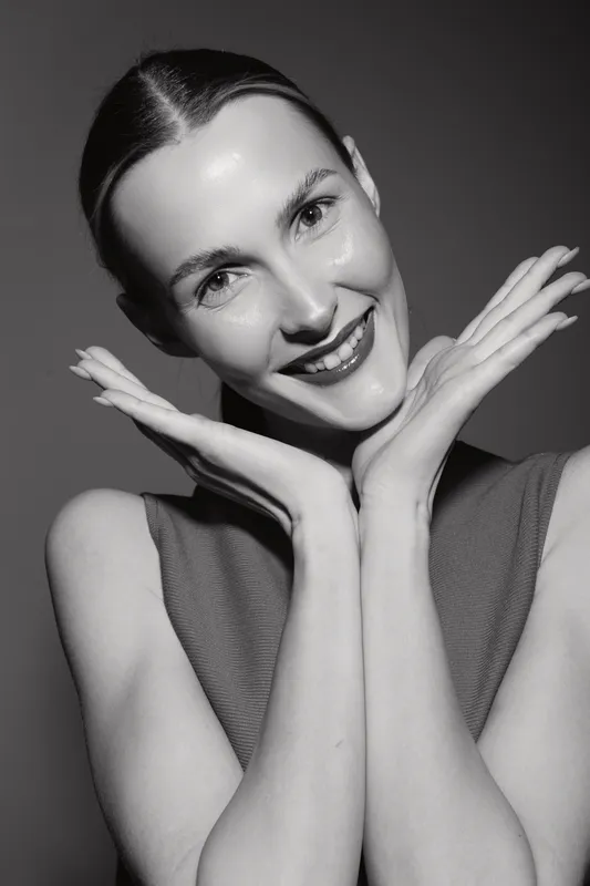 Portrait of a smiling white woman with fair skin and slicked back hair posing with hands near face in studio.