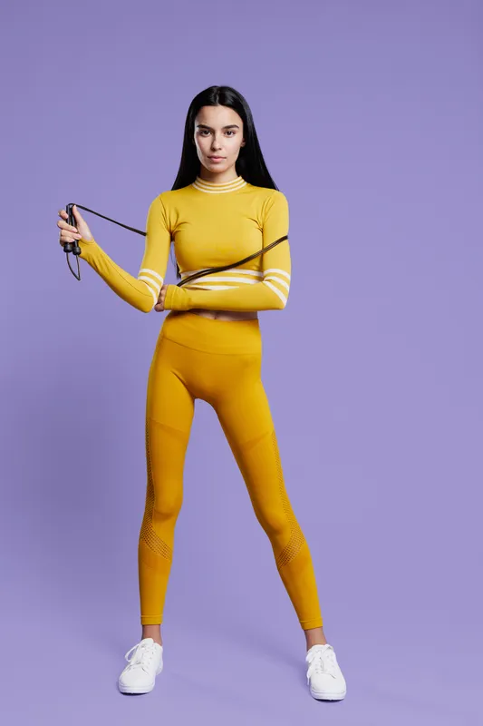 Woman sport fitness dressed in yellow activewear holds a jump rope standing confidently against a purple background, showcasing strength and focus during workout training.