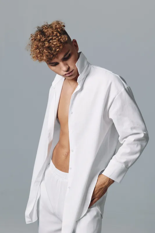 Young man with curly hair wearing white open shirt and pants posing with hand in pocket against gray background in studio for fashion and style concept.