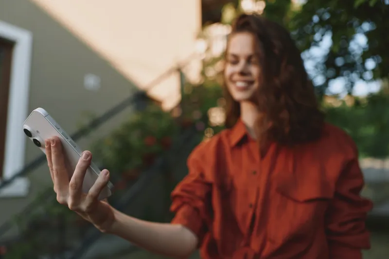 Smiling woman in a red shirt taking a selfie outdoors, capturing a casual lifestyle moment. Natural light creates a warm, friendly mood and carefree vibe