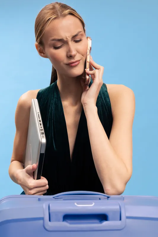 Frustrated woman holding laptop and talking on phone with confused expression. Blue background and suitcase in foreground. Concept of travel stress, technology, and communication issues.