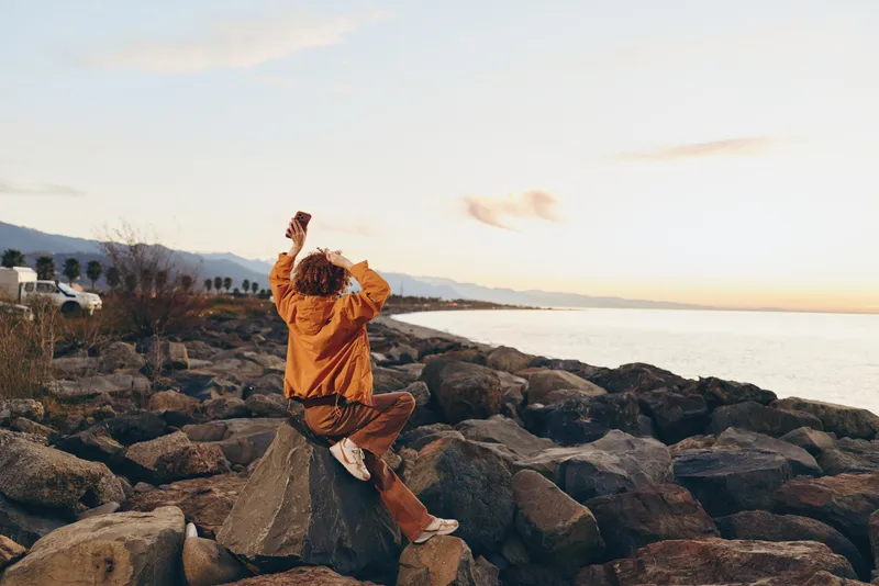 Woman lifestyle sunset ocean rocks outdoors posing casual joy travel, she stands on seaside stones with arms raised, wearing warm coat and scarf, golden sky light, natural smile today
