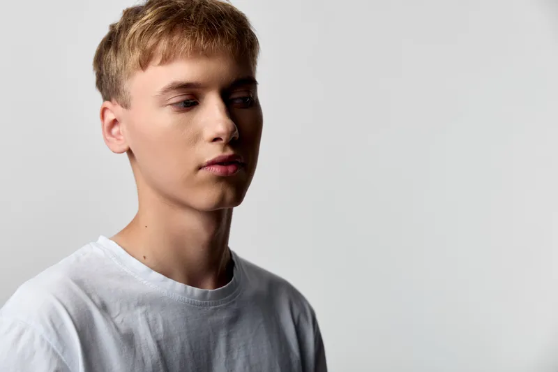 Young man with a calm neutral expression wearing a white t shirt, photographed in studio lighting against a plain background, side view, minimal composition and clean lines for lifestyle or fashion