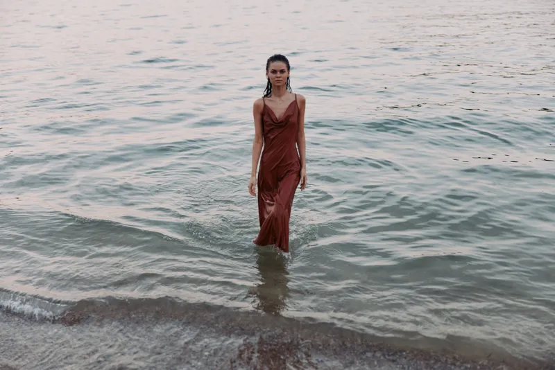 A lone woman in a rust red slip dress stands waist-deep in calm sea water, creating a cinematic, tranquil focal point with soft natural light.