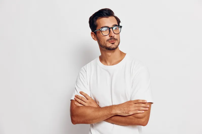 Thoughtful young man in casual white t-shirt and glasses standing with arms crossed against a solid background, exuding a sense of contemplation and style.