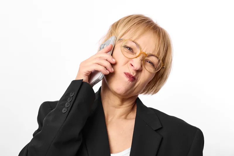 Woman with glasses, phone call communication, stress and tiredness, close portrait in office setting, black blazer, hand near ear, professional indoor neutral white background, concerned frown, soft