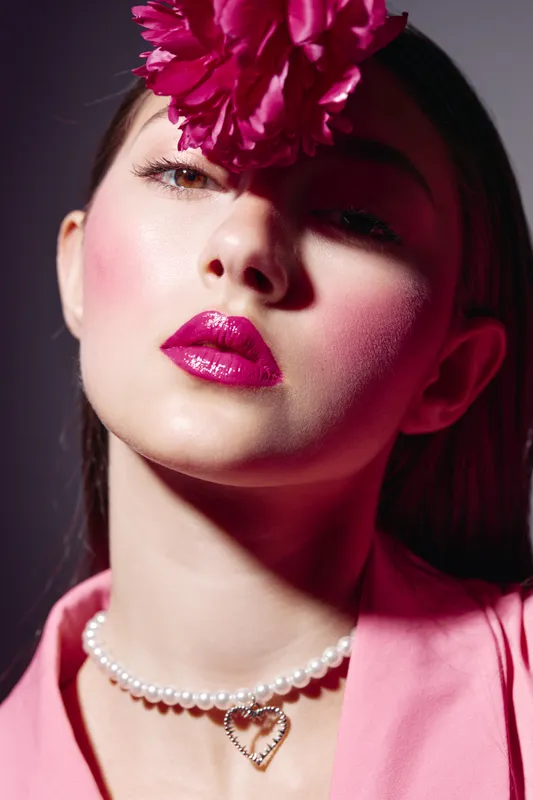 Portrait of a stylish woman with bright makeup and a large flower on her forehead. She wears a pearl necklace and pink outfit. Creative beauty and high fashion concept.
